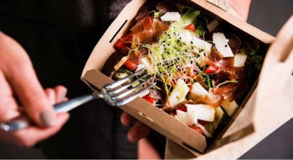 Someone in one hand holding a open takeout box with salad inside and in the other a fork.