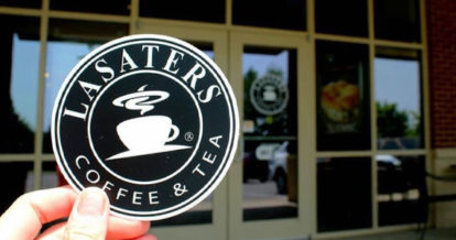 A hand holding up a Lasaters Coffee coaster in front of the coffee shop entrance