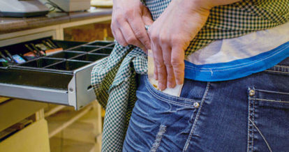 A cashier in front of a open cash drawer putting money in their pocket.