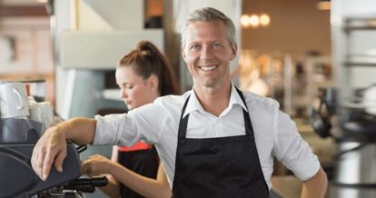 Smiling owner leaning on espresso machine behind cafe counter