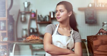 A woman with an apron in her café with her hands crossed looking to the left.