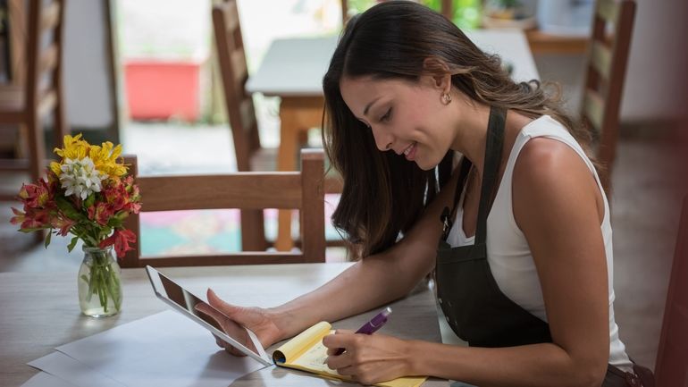 Woman wearing apron, sitting at dining table, holding iPad writing notes on pad of paper.