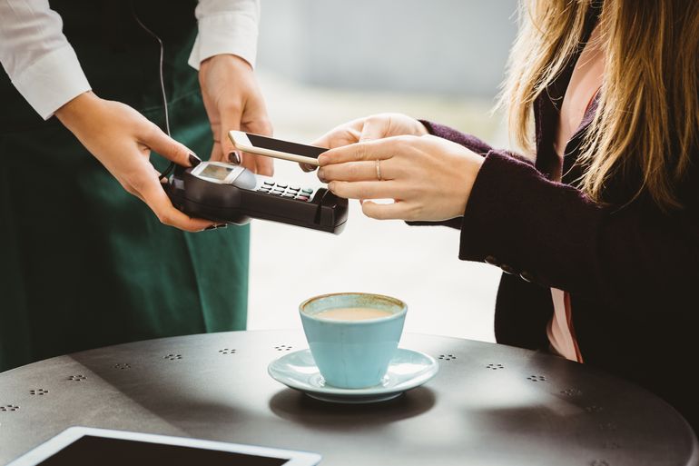 A woman sitting at a table with coffee pays with contactless EMV payment, holding her phone over the terminal