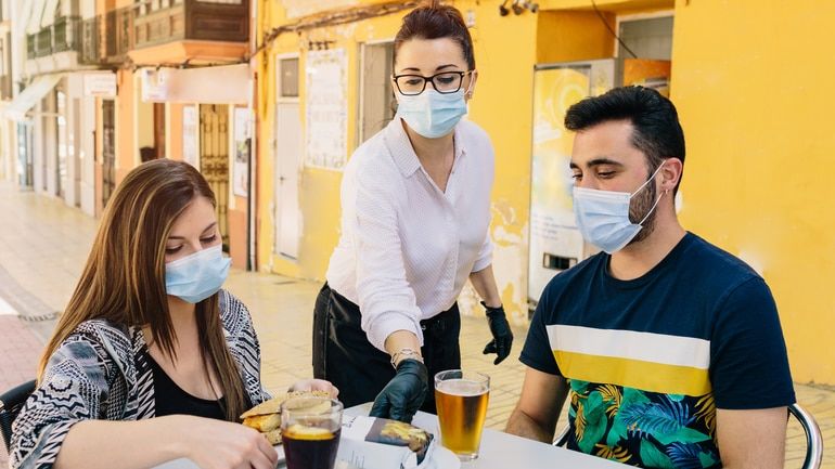 Customers with masks on sitting on the terrace of a bar while attended by a waiter with gloves and masks