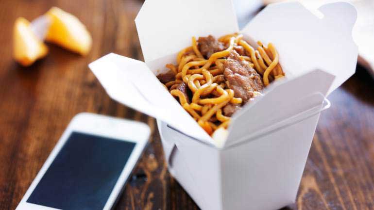 A takeout box of noodles next to a phone