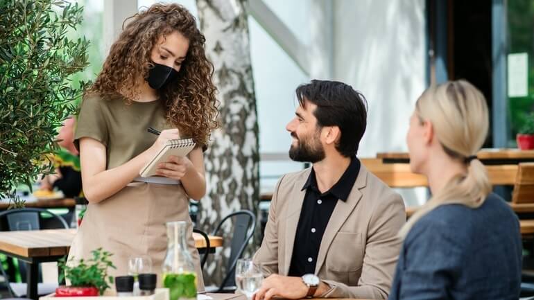 Waitress with face mask serving a couple outdoors on terrace restaurant