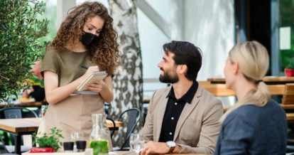 Server tending to a couple at a restaurant patio while wearing a mask and writing down an order on a notepad