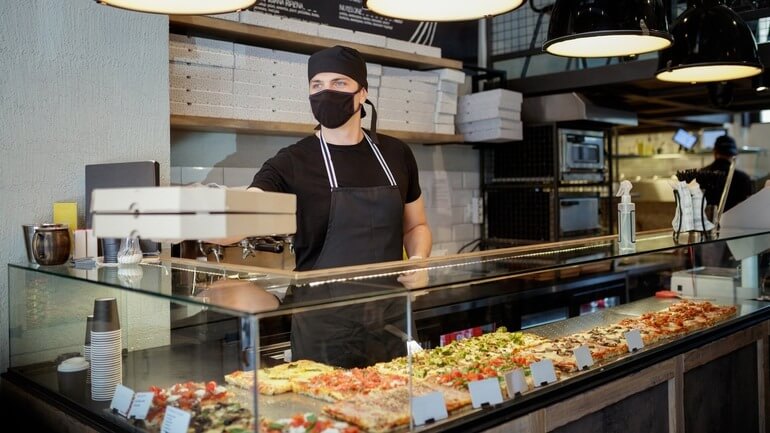 Man in protective mask and gloves passing ordered pizza in boxes over a glass counter