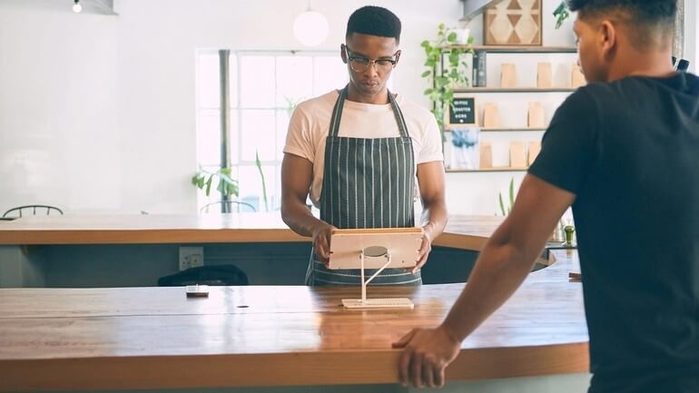 A young man using a digital POS tablet while a customer buys a coffee in a cafe.