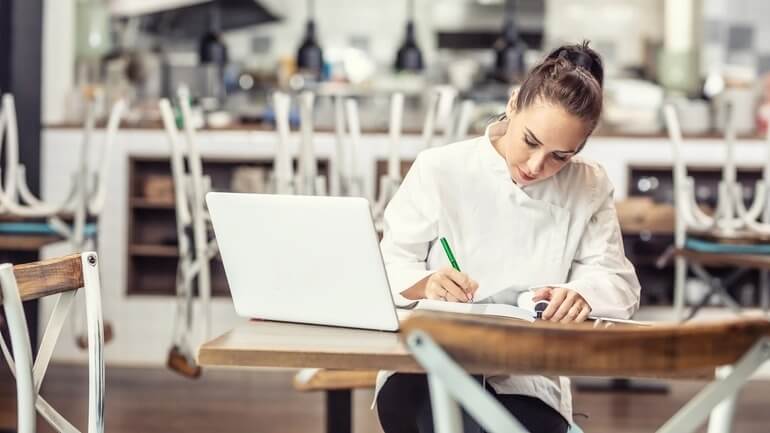 Women in a restaurant writes in a book with a pen and sits in front of her laptop computer at a desk to create a restaurant opening and closing checklist.