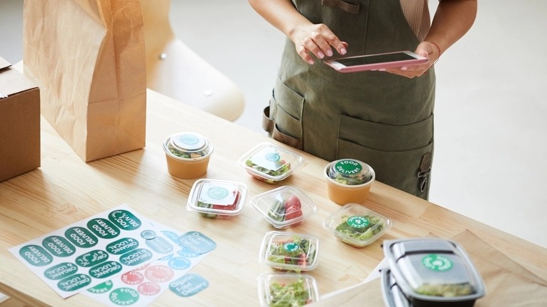 woman plays on tablet while salads in containers and different colors of labels are sitting on a table in front of her