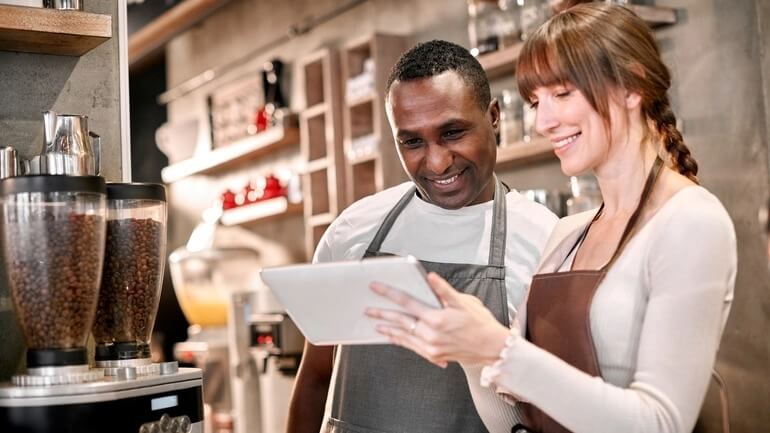 A female manager trains a new male hire as they both read from a handheld tablet in a restaurant.