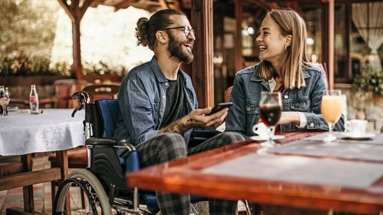 Man in a wheelchair and woman at a restaurant patio enjoying a drink together.