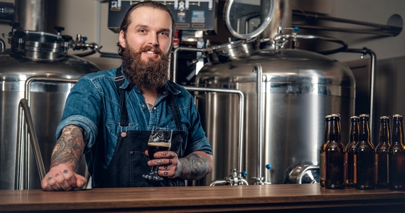 A smiling head brewer holding a beer in front of brewery equipment.