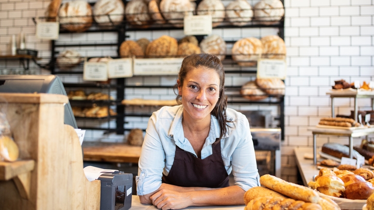 woman looks proud at her bakery surrounded by bread and baked products.