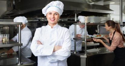 Female chef wearing a white chef's uniform while standing in a commercial restaurant kitchen.