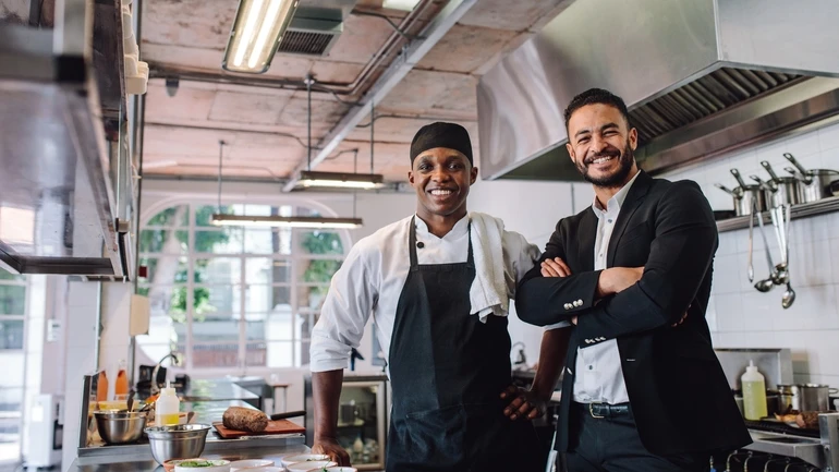 Two restaurant male workers smile at the camera in the kitchen of a restaurant.