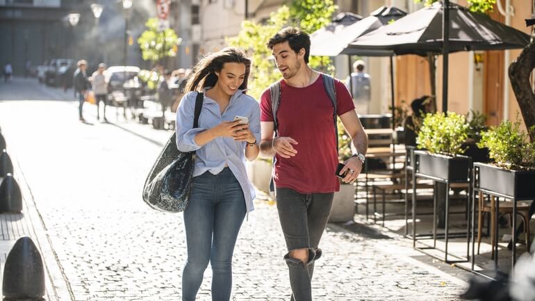 A man and woman stroll down a street filled with cafes while looking at a mobile phone.
