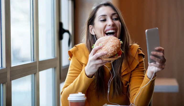 Woman eating a burger while looking at her cell phone and smiling.