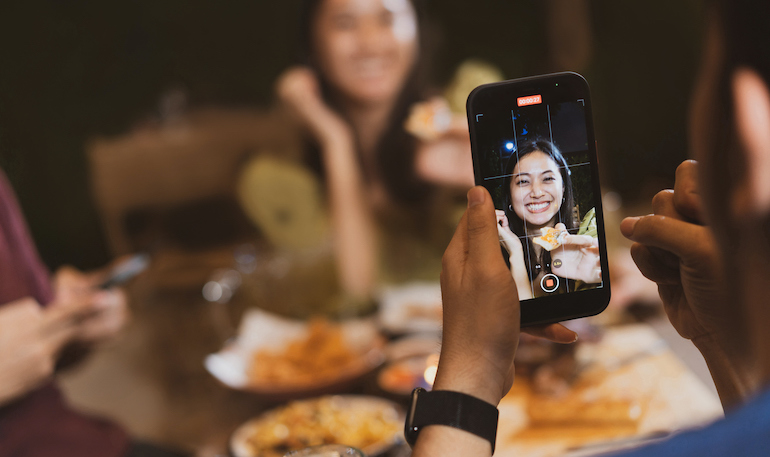 A man taking a video of a woman enjoying food at a restaurant.