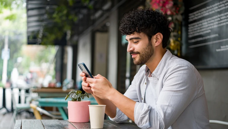 Portrait of a happy Latin American man drinking checking his cell phone at a coffee shop while drinking a cappuccino – lifestyle concepts