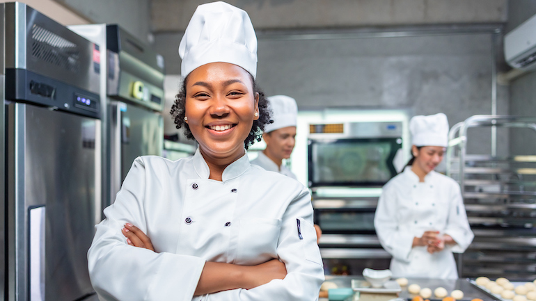 Smiling African female bakers looking at camera. Chefs baker in a chef dress and hat, cooking together in kitchen. Team of professional cooks in uniform preparing meals for a restaurant in the kitchen.