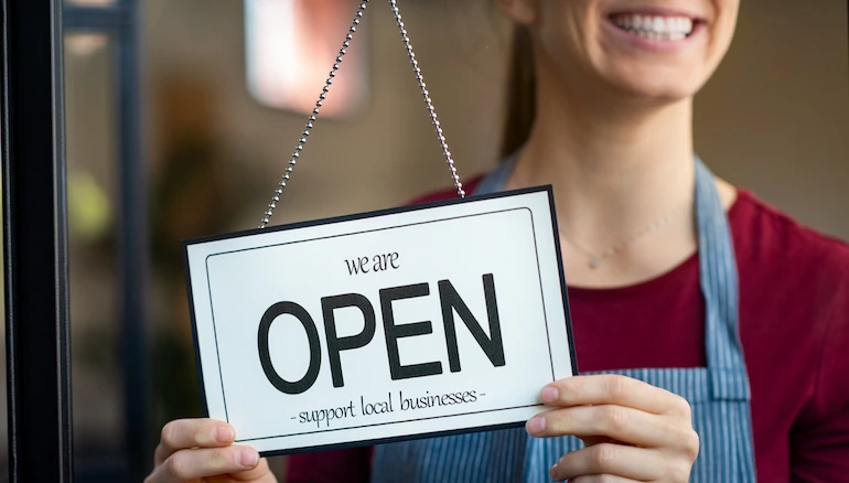 Small business owner smiling while turning the sign for the reopening of the place after the quarantine due to covid-19. Happy businesswoman standing at her restaurant or coffee shop gate with open signboard. Close up of woman’s hands holding sign now we are open support local business.