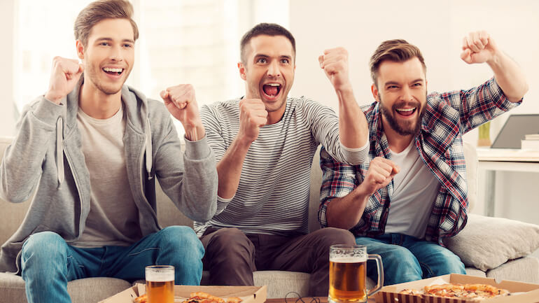 Domestic fans. Three happy young men watching football game and keeping arms raised while sitting on sofa.