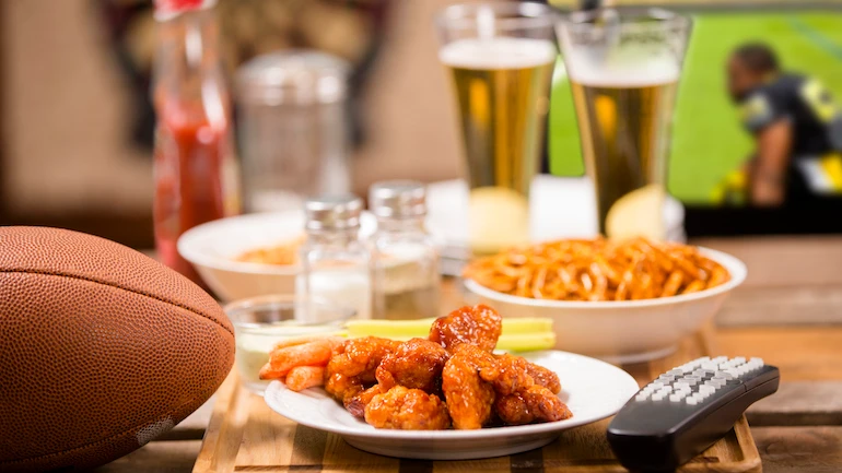 Hot wings and football in foreground.  Beer in mugs in background with television.  Football game on TV in a local pub or sports bar.  Dartboard in background.  Bar top. championship game party!