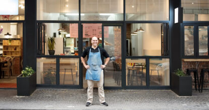 Portrait of confident male restaurant owner standing with his hands on hips outside the restaurant.