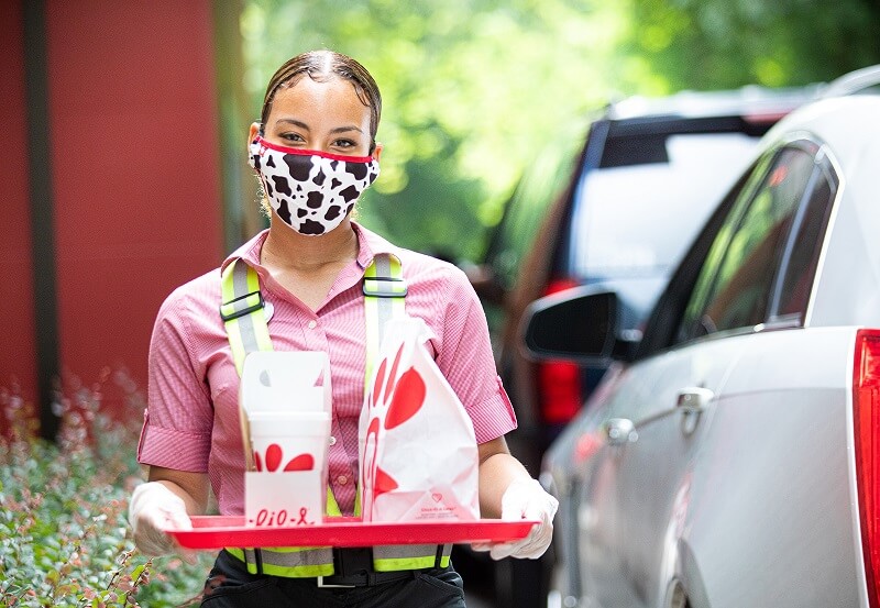 A Chick-fil-A employee wearing a mask and carrying a tray of takeout food through a drive-thru lane.
