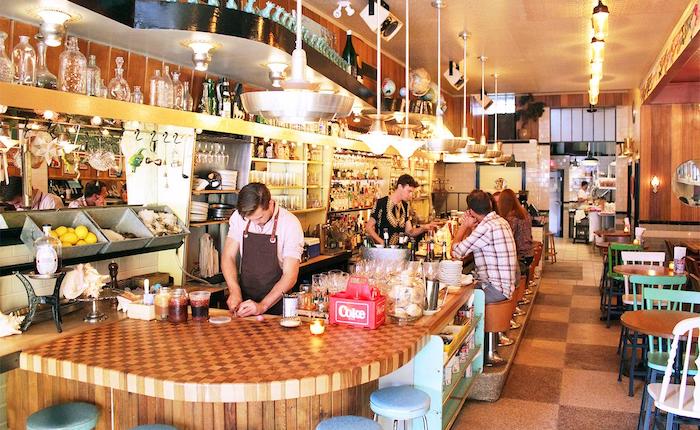 A kitchy Montreal bar with butcher block bar tops, checkered floor tiles, and pink and green chairs.