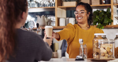 A woman smiling while serving a customer a takeout ccup of coffee in a cafe.
