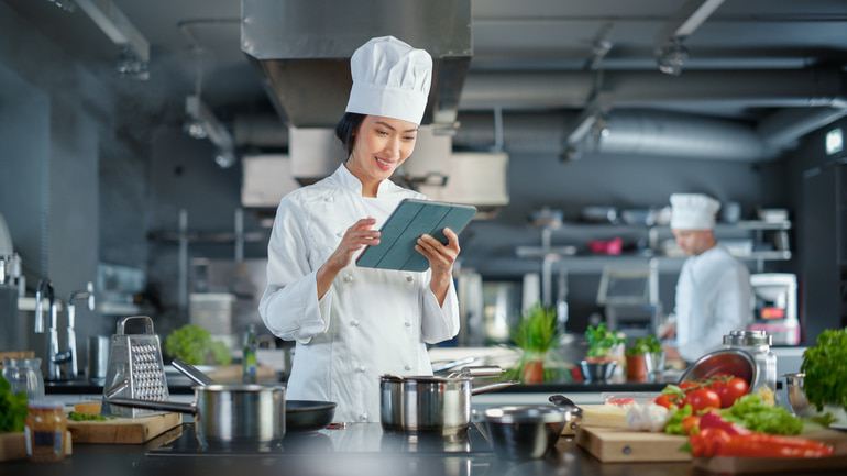 Woman standing in a restaurant kitchen wearing a white chef hat and uniform while holding an iPad.