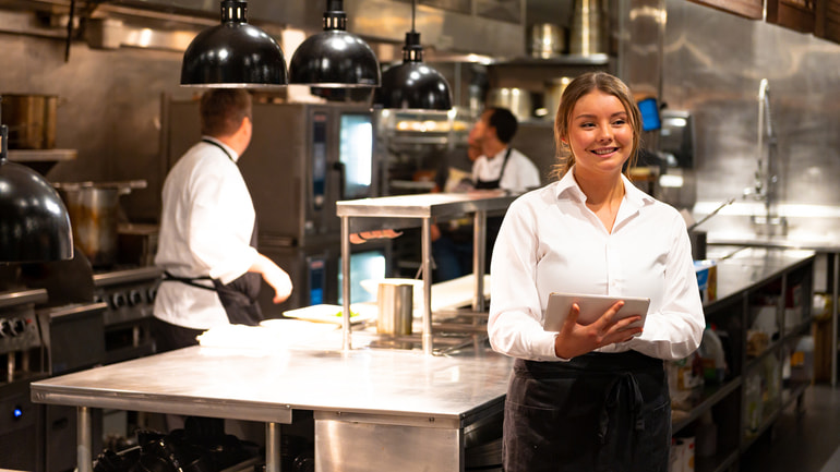 Woman standing smiling in a restaurant kitchen wearing a white dress shirt and black apron.