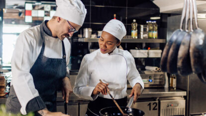 A man and woman wearing a white chef hat and uniform cooking togther in a restaurant kitchen.