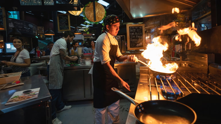 A man wearing a black apron is cooking in front of a restaurant kitchen stove, and there is fire in the wok.