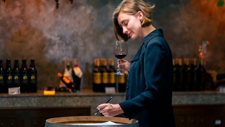 Woman standing holding a glass of wine in one hand and a pen in the other.