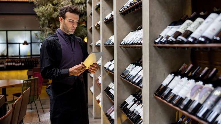 Man wearing a black and purple uniform holding an iPad in front of shelves of wine bottles.