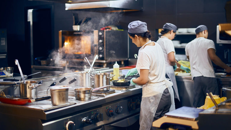 Three chefs wearing white t-shirts and blue hats are in a kitchen cooking.