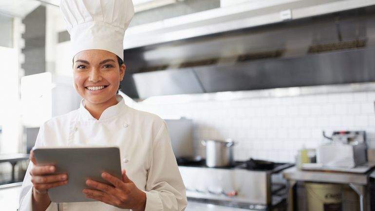 A woman standing and smiling in a kitchen wearing a white chef hat and uniform while holding an iPad.