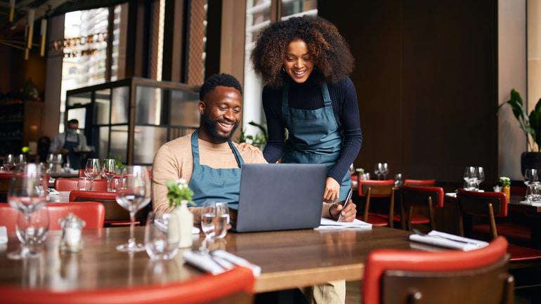 A man sitting in a restaurant dining area in front of a laptop wearing a blue apron with a woman standing next to him also in a blue apron. 