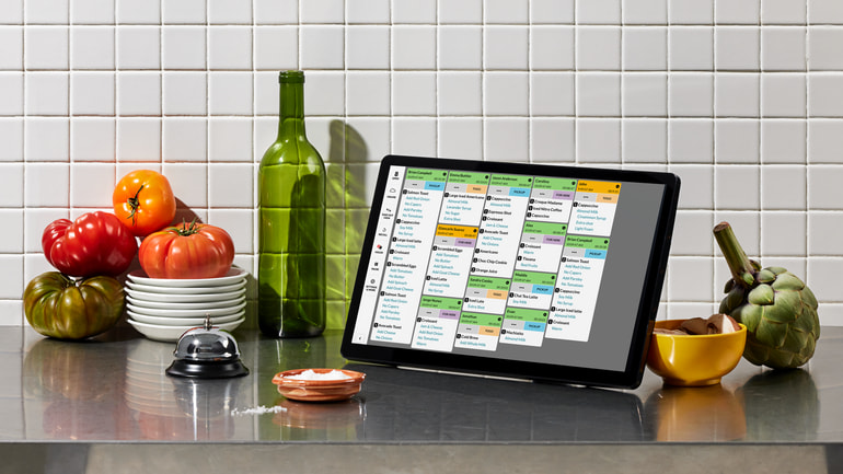 TouchBistro's kitchen display system on a countertop next to a green bottle, silver bell, red tomatoes, and stack of white plates.