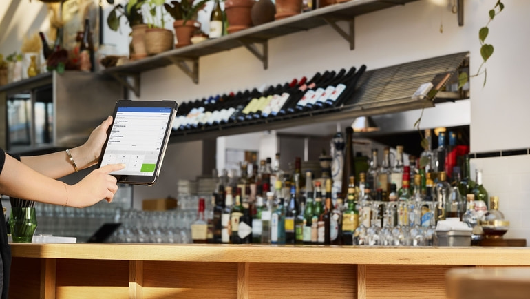A restaurant worker holding a tablet using TouchBistro's profit management software, while standing next to a tanned colored counter with several bottles of alcohol displayed.