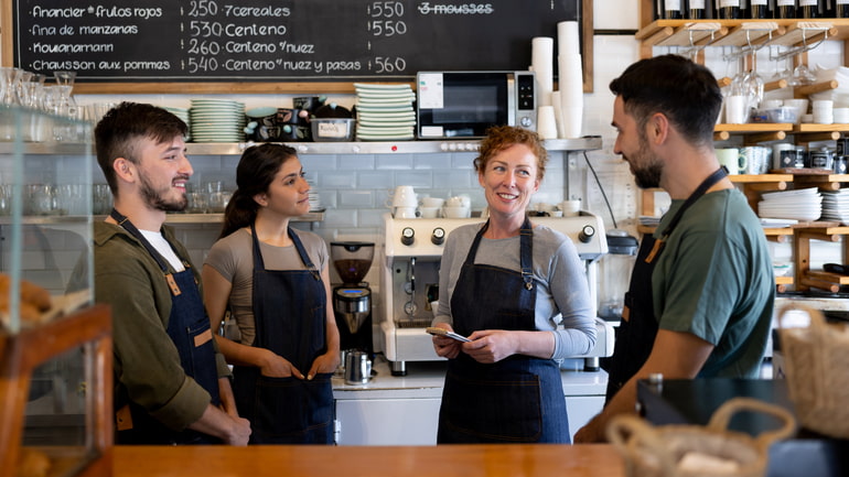 A group of two men and two women who work at a cafe are standing behind the counter talking.