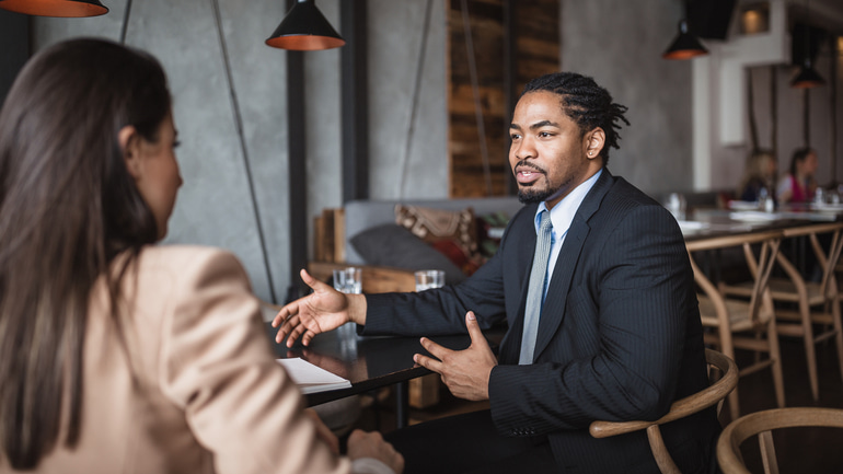 A man wearing a blue dress shirt and black blazer talking to a woman wearing a tan blazer while sitting at a black table in a restaurant.