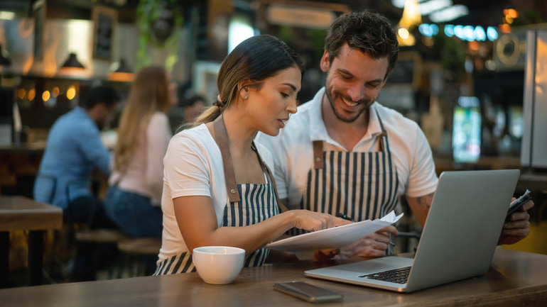 Woman and man wearing white t-shirts and striped aprons are sitting in front of a laptop reading something on a paper.