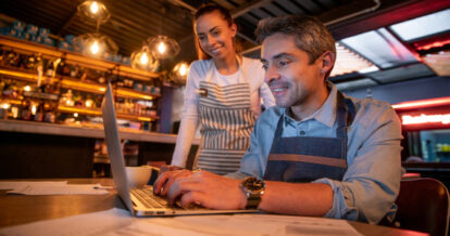 Man sitting in a restaurant doing work on a computer while a woman is standing behind him helping.