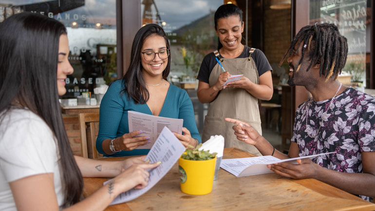 Group of three friends sitting at a restaurant getting their orders taken by a server.