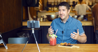 A man filming a video of himself with food and drinks in front of him on his phone.